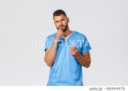 Healthcare workers, medicine, covid-19 and pandemic self-quarantine concept. Serious and thoughtful smart male nurse, doctor in blue scrubs, rub beard and look interested, grey background 66944479