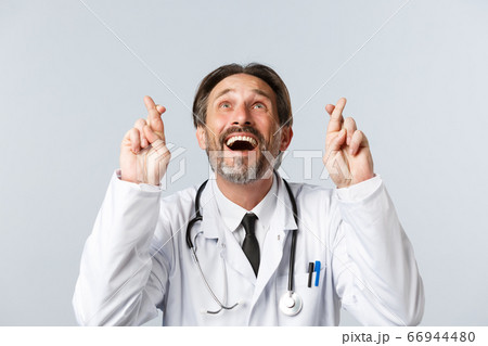 Covid-19, preventing virus, healthcare workers and vaccination concept. Close-up of optimistic hopeful doctor in white coat, cross fingers good luck and looking up smiling, making wish 66944480