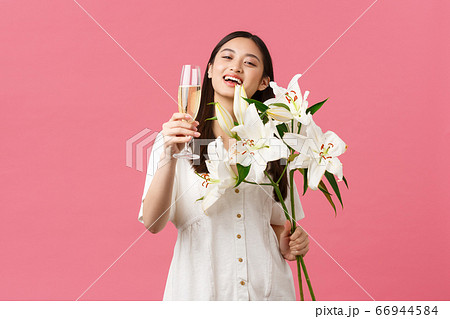 Celebration, party holidays and fun concept. Smiling pretty glamour asian woman in dress with white lilies bouquet, raising glass of champagne to make toast, drink for birthday girl 66944584