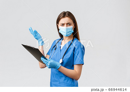 Covid-19, preventing virus, health, healthcare workers and quarantine concept. Displeased and bothered female nurse or doctor in blue scrubs and medical mask, reading nonsense on clipboard 66944614