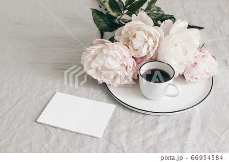 Spring breakfast still life scene. Cup of coffee and bouquet of white and pink peonies on porcelain plate. Blank greeting card mockup on linen table cloth background. Vintage feminine photo. No people 66954584