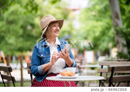 Senior woman with hat sitting outdoors in cafe, using smartphone. 66962808