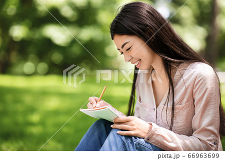 Young asian student girl sitting on lawn in park and writing notes Young asian student girl sitting on lawn in park and writing notes 66963699