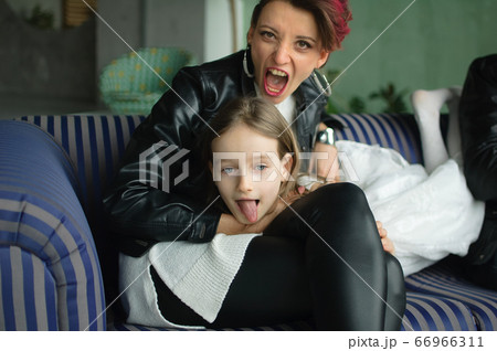 Crazy mother in black leather jaket and pants hugging her little daughter in white dress, they are looking at the camera with opened mouths and grimacing in loft room with houseplants. Crazy mother in black leather jaket and pants hugging her little daughter in white dress, they are looking at the camera with opened mouths and grimacing in loft room with houseplants. 66966311