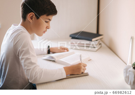 Young boy sitting at desk read the book and write down in notebook. Study at home during quarantine Young boy sitting at desk read the book and write down in notebook. Study at home during quarantine 66982612