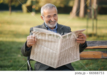 Bearded elderly man in glasses reading newspaper in the autumn park. Handsome gray-haired man sitting on the bench early in the morning Bearded elderly man in glasses reading newspaper in the autumn park. Handsome gray-haired man sitting on the bench early in the morning 66988753