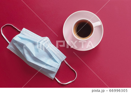 cup of coffee and a removed medical mask. Breakfast in the cafe. Modern reality and life concept. Blue surgical protective mask and pink cup on a red background. cup of coffee and a removed medical mask. Breakfast in the cafe. Modern reality and life concept. Blue surgical protective mask and pink cup on a red background. 66996305