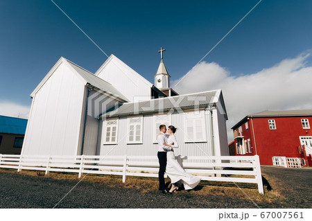 Destination Iceland wedding. Wedding couple near a wooden black church. The groom hugs the bride. White dress of the bride flutters in the wind. Destination Iceland wedding. Wedding couple near a wooden black church. The groom hugs the bride. White dress of the bride flutters in the wind. 67007561