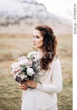 Portrait of a bride in a white wedding dress, with a bride's bouquet in her hands. In a field of dry yellow grass, amid a snowy mountain and grazing Icelandic horses. Destination Iceland wedding.  67008554