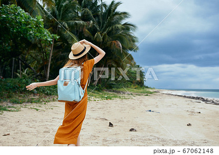 A happy traveler on the island holds a hat on his head and a backpack  67062148