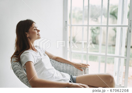 A woman in a T-shirt and shorts sits in a chair near the window A woman in a T-shirt and shorts sits in a chair near the window 67062208