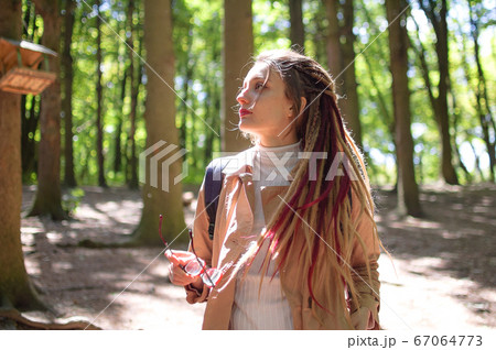 Young woman wearing glasses, casual nude trench, and dreadlocks is standing at the urban park on trees background during sunny spring day outdoors Young woman wearing glasses, casual nude trench, and dreadlocks is standing at the urban park on trees background during sunny spring day outdoors 67064773