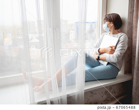 Woman in medical mask had lost her job. She sits on window sill with closed laptop. Lockdown quarantine because of coronavirus COVID19. Self isolation at home. Woman in medical mask had lost her job. She sits on window sill with closed laptop. Lockdown quarantine because of coronavirus COVID19. Self isolation at home. 67065488
