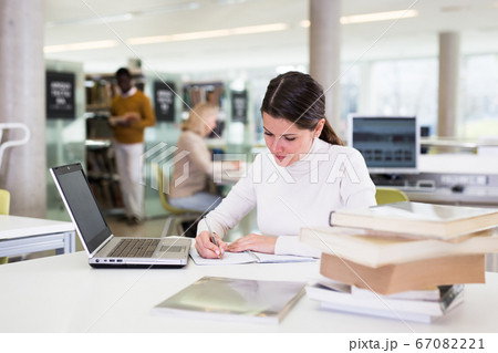 Portrait of smiling woman with laptop and book in public library 67082221