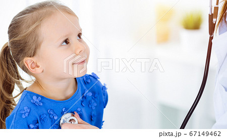 Doctor examining a child by stethoscope in sunny clinic. Happy smiling girl patient dressed in blue dress is at usual medical inspection 67149462
