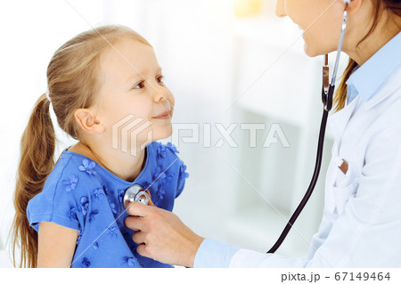 Doctor examining a child by stethoscope in sunny clinic. Happy smiling girl patient dressed in blue dress is at usual medical inspection 67149464