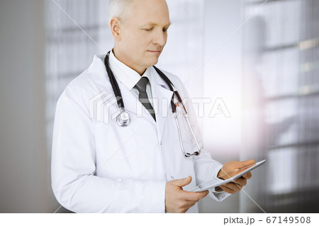 Elderly confident doctor standing straight and using tablet in sunny clinic. Perfect medical service concept Elderly confident doctor standing straight and using tablet in sunny clinic. Perfect medical service concept 67149508