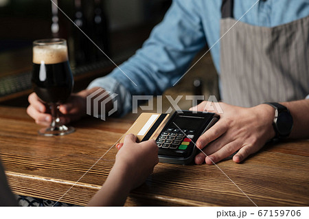 Bar business. Barman in apron gives glass of beer and terminal, girl pays bill with card Bar business. Barman in apron gives glass of beer and terminal, girl pays bill with card 67159706
