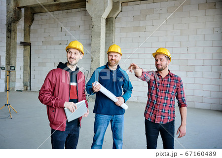 Happy young engineer in hardhat pointing at one of walls of unfinished building Happy young engineer in hardhat pointing at one of walls of unfinished building 67160489