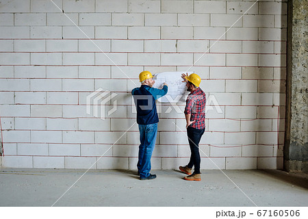 Two professional engineers in hardhats standing by wall and discussing sketch Two professional engineers in hardhats standing by wall and discussing sketch 67160506