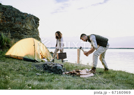 Young male traveler with ax chopping wood on riverbank covered by green grass 67160969