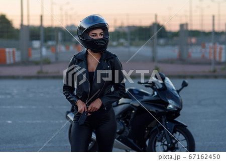 Biker woman in black leather jacket and full face helmet stands near stylish sports motorcycle. Urban parking, sunset in big city. Traveling and active hipster lifestyle. Girls power. 67162450