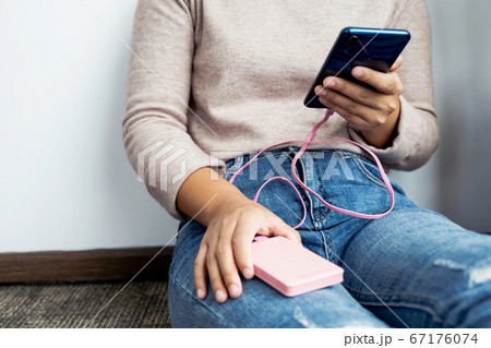 young woman sitting against a wall holding a smartphone while charging a smartphone battery young woman sitting against a wall holding a smartphone while charging a smartphone battery 67176074