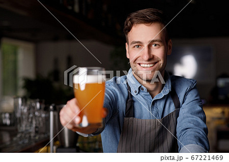 Work in pub. Smiling handsome barman in apron holds out glass of beer Work in pub. Smiling handsome barman in apron holds out glass of beer 67221469