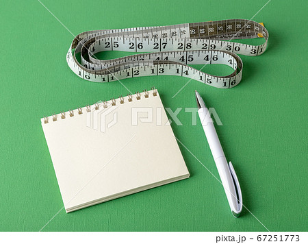 Measuring tape with centimeters and inches, paper notepad and white pen on a green background. 67251773