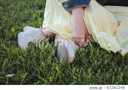 A girl in white sports sneakers and a yellow skirt sits on the grass in the park. Legs and hand closeup A girl in white sports sneakers and a yellow skirt sits on the grass in the park. Legs and hand closeup 67261246