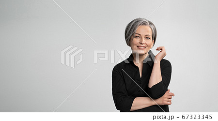 Mature Caucasian woman smiling charmingly with hand near face. Graying elegant woman with short bob hairstyle looks at camera posing on white background. Blank or template with text space at left 67323345