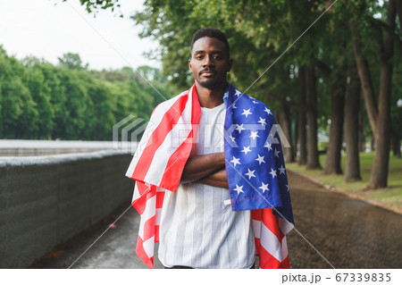 Afro American man with USA flag on his shoulders looking at camera, standing outdoors. Day summer Afro American man with USA flag on his shoulders looking at camera, standing outdoors. Day summer 67339835