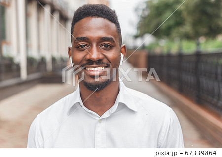 Portrait of a smiling black businessman with wireless earphones walking outdoors 67339864