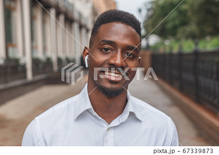 Portrait of a smiling black businessman with wireless earphones walking outdoors 67339873