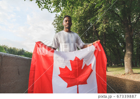 Handsome Afro American man with Canadian flag seriously looking at camera, standing outdoors. 67339887