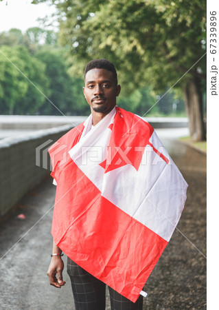 Handsome Afro American man with Canadian flag on his shoulders seriously looking at camera, standing outdoors. 67339896