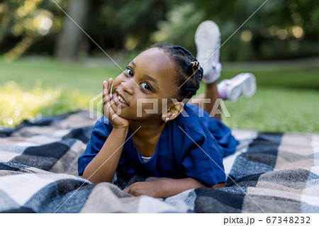 Outdoor portrait of a cute little African girl lying down on the checkered blanket in the park and smiling. Adorable child having fun on a walk Outdoor portrait of a cute little African girl lying down on the checkered blanket in the park and smiling. Adorable child having fun on a walk 67348232