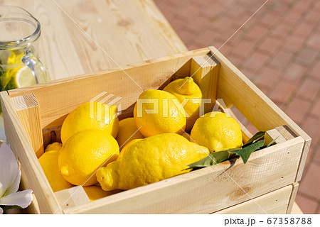 Pile of fresh lemons in wooden box standing by glass jug with citrus slices Pile of fresh lemons in wooden box standing by glass jug with citrus slices 67358788