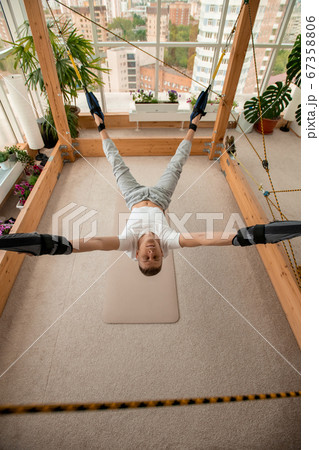 Young sportsman in activewear hanging over the floor during aerial yoga workout 67358806