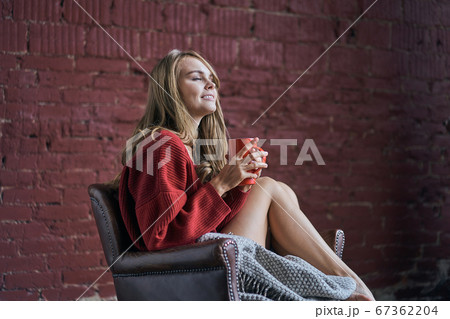 Young woman with closed eyes holding red cup of coffee or tea drinking warm beverage sitting in cozy chair at loft apartment. Happy healthy lady relaxing with morning drink in hands enjoying peace of Young woman with closed eyes holding red cup of coffee or tea drinking warm beverage sitting in cozy chair at loft apartment. Happy healthy lady relaxing with morning drink in hands enjoying peace of 67362204
