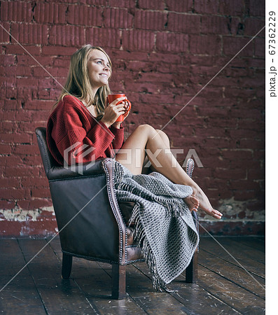 Young smiling woman holding red cup of coffee or tea drinking warm beverage sitting in cozy chair at loft apartment. Happy healthy lady relaxing with morning drink in hands enjoying peace of mind. Young smiling woman holding red cup of coffee or tea drinking warm beverage sitting in cozy chair at loft apartment. Happy healthy lady relaxing with morning drink in hands enjoying peace of mind. 67362229