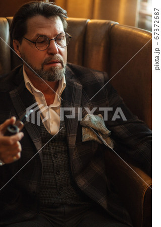 Reflective well-dressed mature bearded man in glasses smoking tobacco pipe Reflective well-dressed mature bearded man in glasses smoking tobacco pipe 67372687