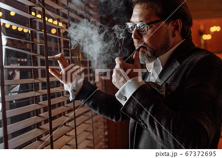 Reflective well-dressed mature bearded man in glasses smoking tobacco pipe 67372695