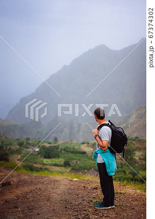Hiker with backpack in arid terrain in front of a incredible panorama view of high mountain ranges deep ravines and coastline. Santo Antao Cape Verde 67374302