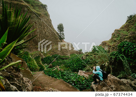 Male hiker walking through a wondrous misty landscape. Huge rocks surround a fertile ravine full of lotus plants. Santo Antao Cape Verde 67374434