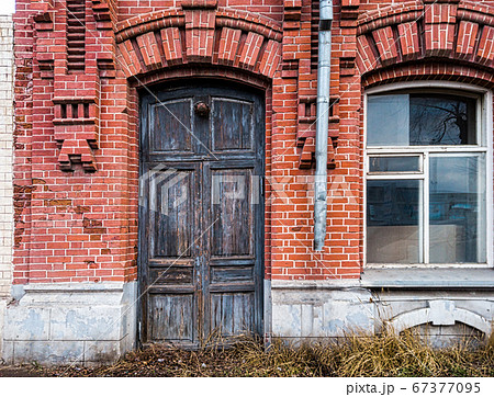 The wooden door in the wall of the old brick building is closed and there is a window nearby 67377095