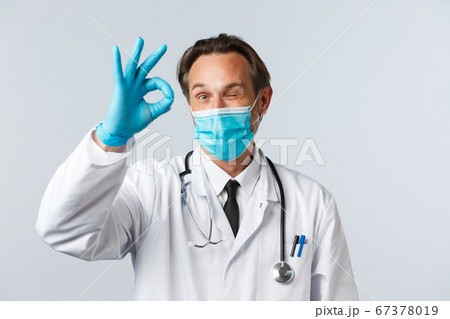 Covid-19, preventing virus, healthcare workers and vaccination concept. Cheerful satisfied doctor in medical mask and gloves show okay sign and wink. Physician give approval, agree with patient Covid-19, preventing virus, healthcare workers and vaccination concept. Cheerful satisfied doctor in medical mask and gloves show okay sign and wink. Physician give approval, agree with patient 67378019