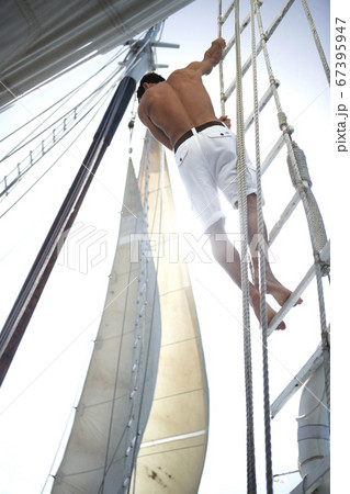 A man in white shorts with bare chest climbing up the rigging of a sailing boat. A man in white shorts with bare chest climbing up the rigging of a sailing boat. 67395947