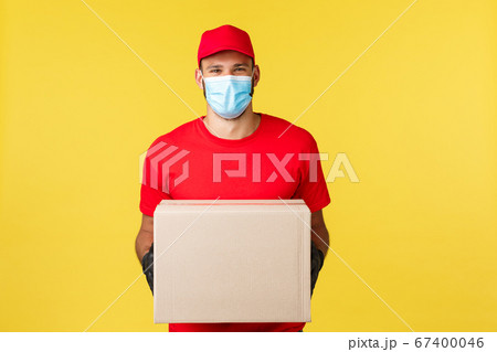 Express delivery during pandemic, covid-19, safe shipping, online shopping concept. Smiling pleasant courier in red uniform, medical mask, provide contactless deliver order, holding package box 67400046