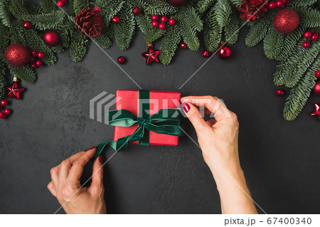 Woman wrapped Xmas red gift with green ribbon on black. Handmade workshop. New Year. Top view, flat lay. Christmas time. 67400340